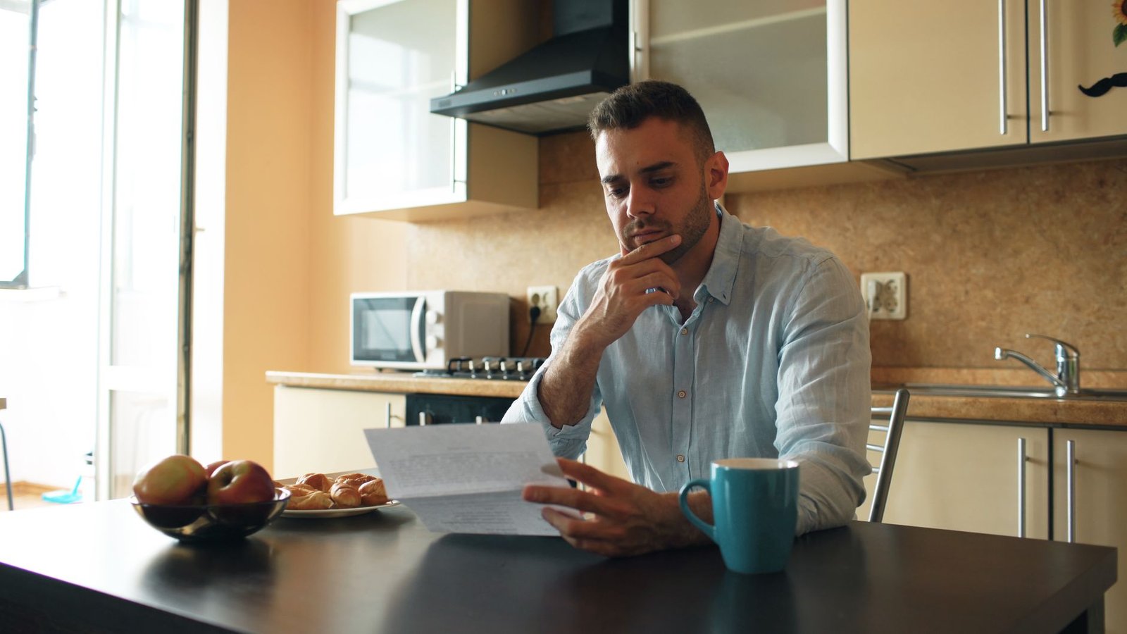 A man reads a letter at a kitchen table in the morning light